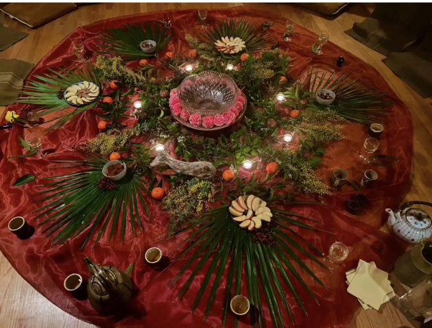 A community altar of flowers, fruit, candles and greenery on a red cloth